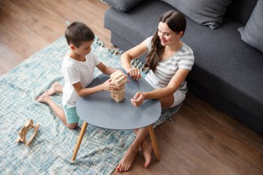 Küçük anaokulu çocuğu, mutlu anneye sarılıp tahta masa oyunu oynuyor, evde güzel vakit geçiriyor..