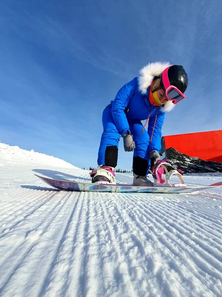 A lány rögzíti a snowboard rögzítéseket. Egy snowboardos lány a havas hegyekben, ül a hóban, rögzíti a hegyet, és készül a leereszkedésre. — Stock Fotó