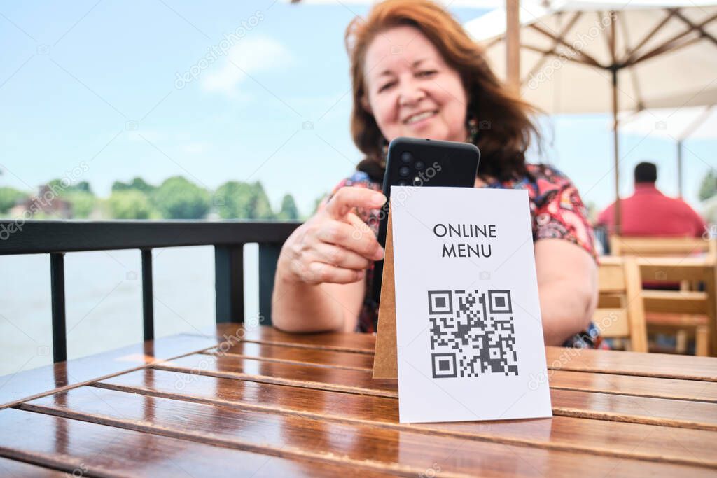 Mature latin woman smiling scanning a QR code with her smartphone to access a restaurant menu; use of contactless technology in everyday life.