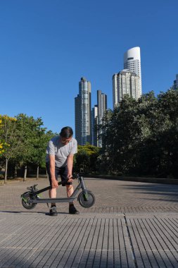 İspanyol son sınıf öğrencisi güneşli bir sabah bir şehir parkında katlanmış bir elektrikli scooter tutuyor. Temiz enerji, yeşil hareketlilik, ekolojik ulaşım ve aktif yaşam kavramları.