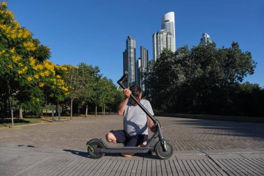 İspanyol kıdemli bir adam güneşli bir sabah bir şehir parkında elektrikli scooterını açıyor. Elektrik enerjisi, yeşil hareketlilik, çevre dostu ulaşım ve aktif kentsel yaşam kavramları.