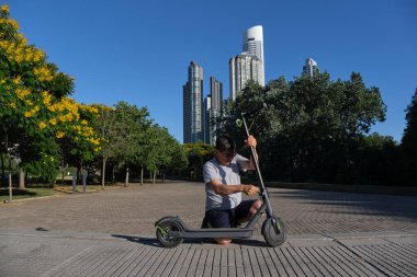 İspanyol kıdemli bir adam güneşli bir sabah bir şehir parkında elektrikli scooterını açıyor. Elektrik enerjisi, yeşil hareketlilik, çevre dostu ulaşım ve aktif kentsel yaşam kavramları.