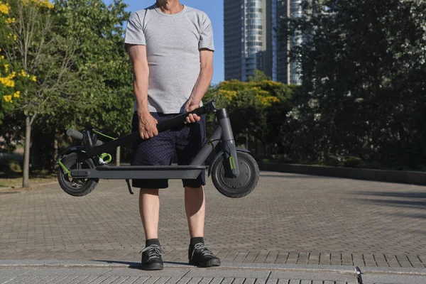 認識できない男は晴れた朝に都市公園で折り畳まれた電動キックスクーターを保持しています クリーンエネルギー グリーンモビリティ エコ輸送 アクティブライフの概念 — ストック写真