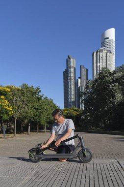 İspanyol kıdemli bir adam güneşli bir sabah bir şehir parkında elektrikli scooterını açıyor. Elektrik enerjisi, yeşil hareketlilik, ekolojik ulaşım ve aktif kentsel yaşam kavramları.