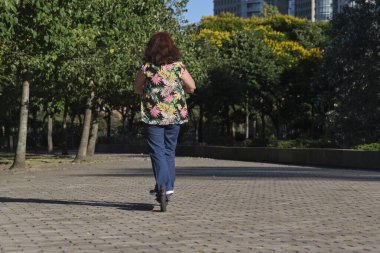 Şehirde güneşli bir sabah, arkadan manzaralı, elektrikli scooterıyla gezen İspanyol olgun bir kadın. Aktif kentsel yaşam, modern hareketlilik ve ekolojik ulaşım kavramları.