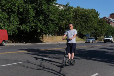 Güneşli bir sabah, İspanyol asıllı son sınıf öğrencisi, şehir caddesinde elektrikli scooterına biniyor. Aktif emeklilik hayatı, modern hareketlilik ve ekolojik taşımacılık kavramları.