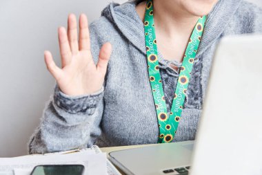 Unrecognizable person using a sunflower lanyard, symbol of people with invisible or hidden disabilities, waves at a laptop camera during a remote class or video call. clipart