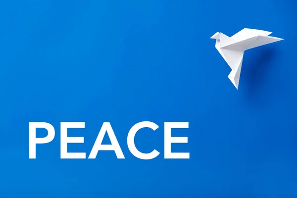 Word Peace in white letters on a blue background next to a flying white paper dove, symbol of peace.
