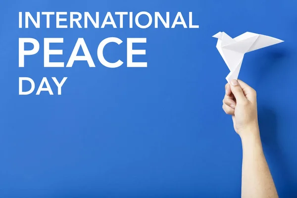 Text International Peace Day in white letters on a blue background next to a hand holding a white paper dove, symbol of peace.
