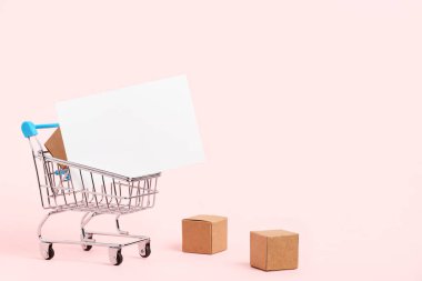 Shopping cart with cardboard boxes and a blank white paper poster on a pastel pink background. Minimalist design with copy space. Concepts: market deals, black friday, logistics and packaging.