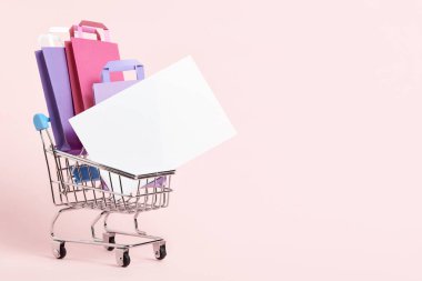 Shopping cart loaded with paper bags and a blank card on a pastel pink background. Minimalist design with copy space. Concepts: market deals, seasonal sales and discounts, black friday.