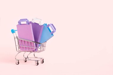 Shopping cart with purple, lilac and blue paper bags on a pastel pink background. Minimalist design with copy space. Concepts: market deals, seasonal discounts and rebates, black friday sales.