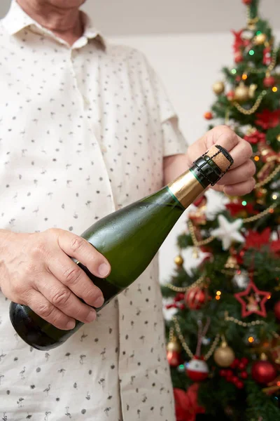 Unrecognizable man opening a bottle of champagne or cider with a Christmas tree in the background. Concept of celebratory toast.