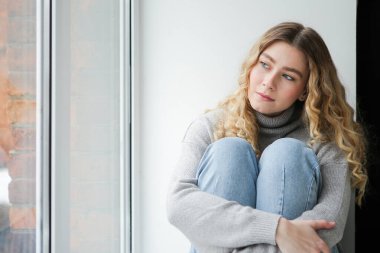 Gri kazaklı ve mavi kotlu bir kadın pencere eşiğinde oturup pencereden izliyor. Depresyondaki kız pencerede düşünüyor