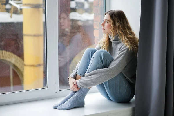 Gri kazaklı ve mavi kotlu bir kadın pencere eşiğinde oturup pencereden izliyor. Depresyondaki kız pencerede düşünüyor