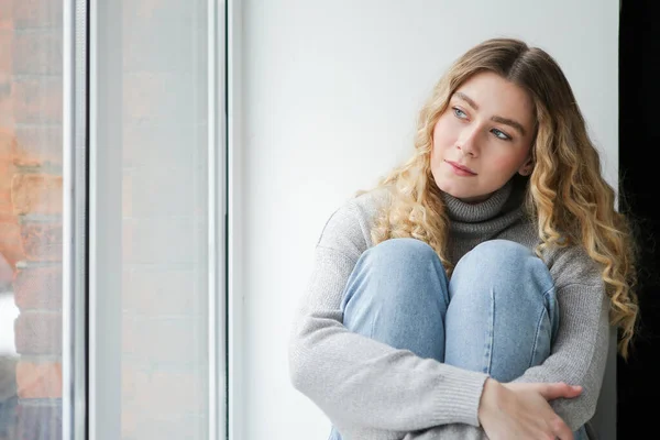 Gri kazaklı ve mavi kotlu bir kadın pencere eşiğinde oturup pencereden izliyor. Depresyondaki kız pencerede düşünüyor