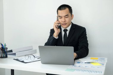 İşadamı patronu akıllı telefon kullanarak müşteriyle yeni iş yatırımları hakkında konuşuyor ve ofiste çalışırken iş projesi verilerini kaydetmek için dizüstü bilgisayarda yazı yazıyor.