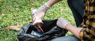 Eldivenli gönüllü bir adam, çevre aktiviteleri sırasında çöpleri toplamak için kahve fincanını plastik siyah bir torbaya koyup parkı temizliyor.