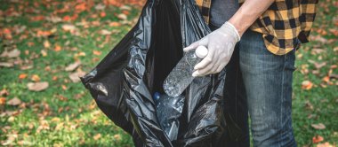 Eldivenli gönüllü bir adam plastik siyah çanta taşımak için yürüyor ve çöp toplamak için çevre aktiviteleri sırasında parkı temizlemek için plastik şişe topluyor.