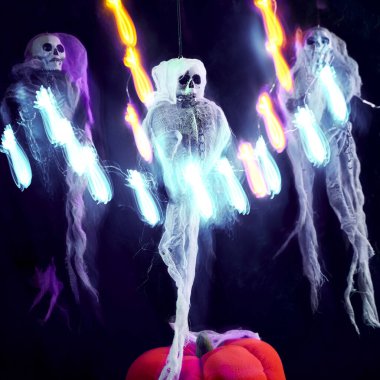 Cadılar Bayramı dekorasyonu. Çok renkli bir pelerinle süslenmiş üç iskelet. Koyu arkaplanda parlayan balkabağı ve örümceklerden oluşan bir çelenkle süslenmiş. Çelenklerin ışığı sanatsal olarak bulanık..