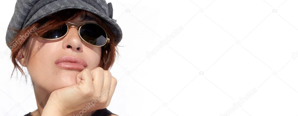 Close-up portrait of a confident young woman with modern wispy hairstyle wearing a fashionable peaked cap and sunglasses resting her chin on hand. Panorama banner with copy space on white background.