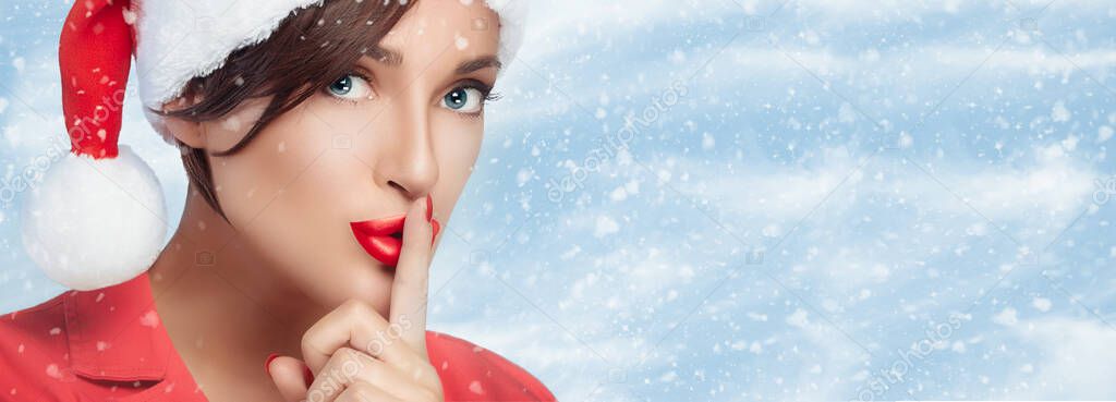 Christmas tale concept banner with the close-up portrait of a beautiful mysterious woman wearing Santa hat while making the shushing sign to keep quiet against winter cloudy sky