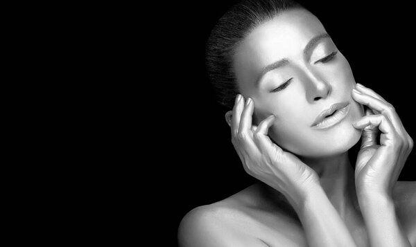 Silver skin model girl. Beautiful sensual young woman with glowing metallic silver body makeup raising hands to her face with eyes closed and a serene expression. Monochrome portrait isolated on black