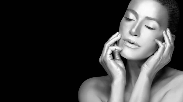 Silver skin model girl. Beautiful sensual young woman with glowing metallic silver body makeup raising hands to her face with eyes closed and a serene expression. Monochrome portrait isolated on black