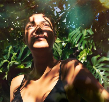 Bikini giymiş, yeşil egzotik bir parkta duran, yüzü ve vücudu gölgeli bir kadın yaz güneşinin keyfini çıkarıyor.