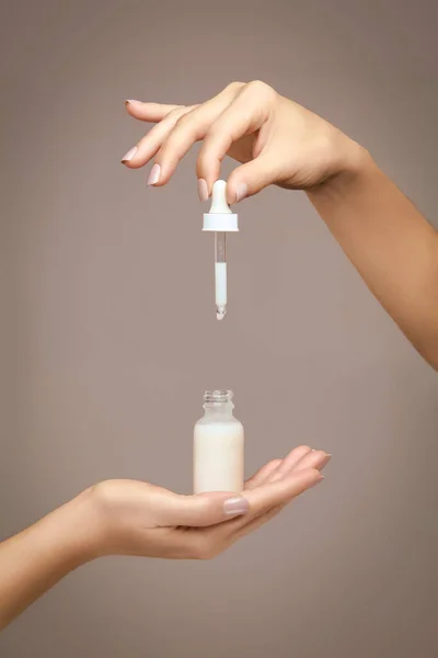 Female hands holding an unlabelled generic dropper bottle filled with white cosmetic fluid over a beige background in a cosmetology and beauty concept