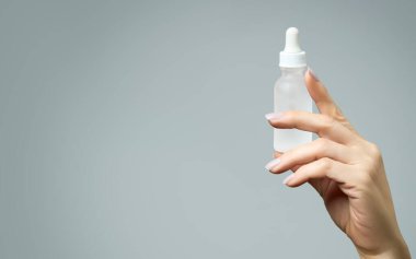 Kadın eli, kozmetoloji ve cilt bakımı konseptinde etiketsiz bir damlatıcıyı tutuyor. Metnin yan tarafında, gri arkaplan üzerinde, metin için kopyalama alanı var.