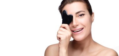 Sağlıklı, orta yaşlı, gülümseyen bir kadın kameraya gülümserken bir Gua sha aleti sergiliyor. Güzellik portresi kopyalama alanı ile beyaza izole edildi