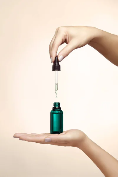 Female hands holding an unlabelled generic dropper bottle filled with cosmetic fluid over a beige background in a cosmetology and beauty concept