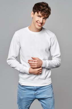 Beyaz kazaklı yakışıklı adam gri arka planda poz veriyor.