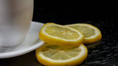 Siyah arka planda çay olan beyaz bardak. Siyah ahşap bir masada limon dilimleri.
