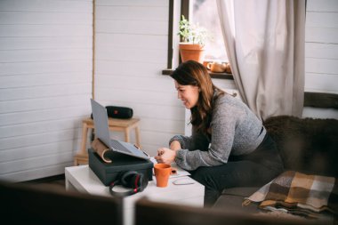 Bir kadın oturma odasında dizüstü bilgisayarla oturuyor, ekrana bakıyor, notlar alıyor, internetten yabancı bir dil öğreniyor, online eğitim kursları okuyor, internetten eğitim alıyor, bir müşteriye danışmanlık yapıyor.