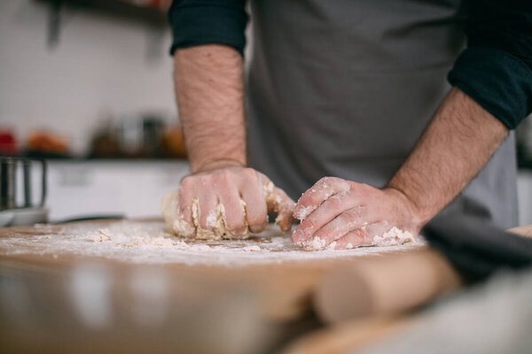 A male chef prepares noodle dough at home in the kitchen. Close up of hands with flour and dough. Young guy in an apron in his kitchen at the house at the table with ingredients for cooking noodles