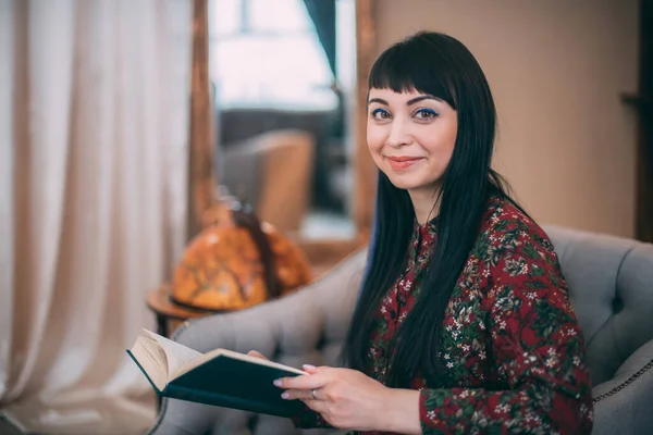 Portrait of a beautiful bright, smart writer with a book in her hands in a cozy living room. Premiere of the book, portrait of the author. A young black-haired woman sits in a chair, holding her book.