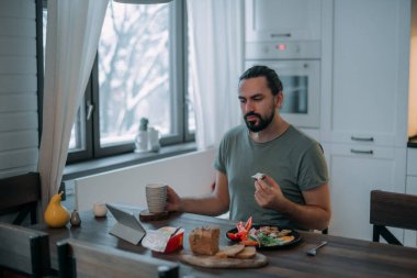 Bir adam evde tabletle kahvaltı yapıyor. Genç yakışıklı beyaz adam yemek yer, kahve içer, sabahın erken saatlerinde büyük, parlak bir mutfakta tablete bakar.