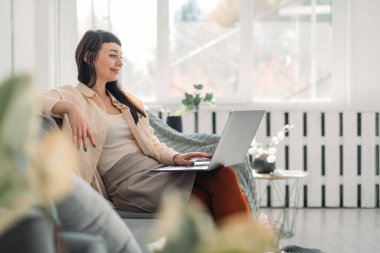 Genç bir kadın dizüstü bilgisayarda çalışıyor, güneşli bir günde evdeki kanepede internetten iletişim kuruyor. Evde kıyafetleri olan esmer bir kadın internette geziniyor, rahat bir koltukta otururken video izliyor.