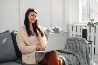 Genç bir kadın dizüstü bilgisayarda çalışıyor, güneşli bir günde modern ve parlak bir ofiste internetten iletişim kuruyor. Güzel esmer kız kanepede oturur, çalışır, video konferansı yönetir, online ders verir.