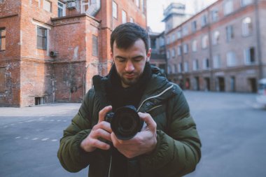 Elinde fotoğraf makinesi olan genç bir erkek fotoğrafçının portresi sokakta, eski bir avluda fotoğraf ya da video çekiyor. Eski bir tuğla binanın arka planında profesyonel kamerası olan yakışıklı bir adam.