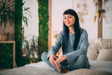 Özel günlüğü olan genç bir kız rahat bir yatak odasında mavi pijamalarıyla yatağın üzerinde. Güzel esmer kadın, düşünceler günlüğüne büyük bir yatağa konforlu ev kıyafetleriyle anılar yazar.