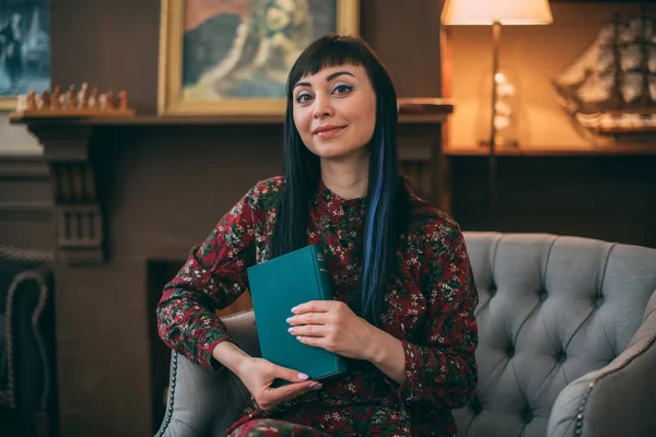 Portrait of a beautiful bright, smart writer with a book in her hands in a cozy living room. Premiere of the book, portrait of the author. A young black-haired woman sits in a chair, holding her book.