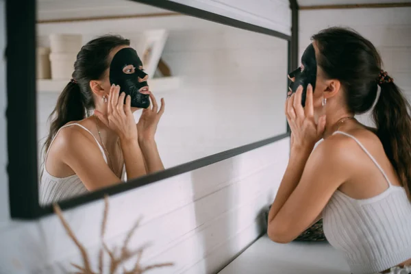 Portrait of a young woman with a black face mask at the mirror at home. A beautiful brunette woman makes facial skin care procedures with a cleansing, moisturizing mask near the mirror in a bright room