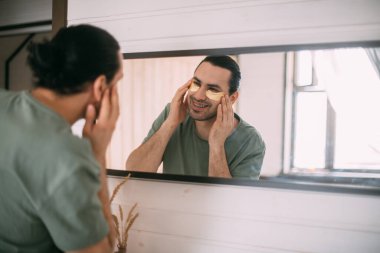 Evde spa günü. Evdeki aynada gözlerinin altında yamalar olan genç bir adam. Yakışıklı bir esmer, parlak bir odadaki büyük bir aynanın yanında hyaluronik yamalarla yüz bakımı prosedürleri uygular..