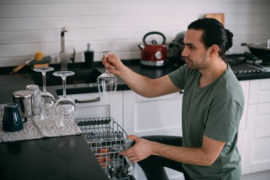 Hafta sonu ödevi. Genç bir adam evdeki mutfaktaki bulaşık makinesinden temiz bulaşıkları çıkarıyor. Ev kıyafetleri içinde yakışıklı bir adam büyük, rahat bir mutfakta bulaşık yıkıyor.