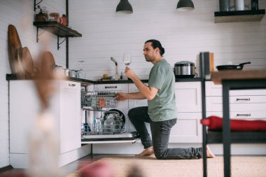 Hafta sonu ödevi. Genç bir adam evdeki mutfaktaki bulaşık makinesinden temiz bulaşıkları çıkarıyor. Ev kıyafetleri içinde yakışıklı bir adam büyük, rahat bir mutfakta bulaşık yıkıyor.