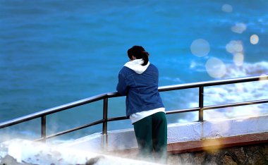 Ceketli genç bir kadın deniz kenarındaki denize bakıyor.