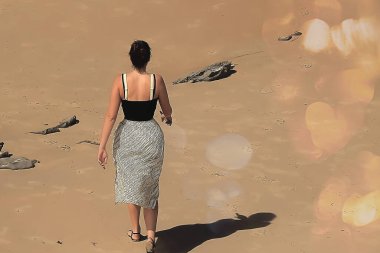 Etekli ve siyah bluzlu genç bir kadın sahilde yürüyor.